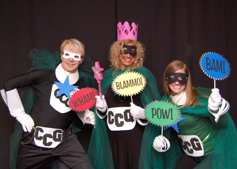One man and two women dressed as superheroes wearing eye masks and holding signs with action sounds.