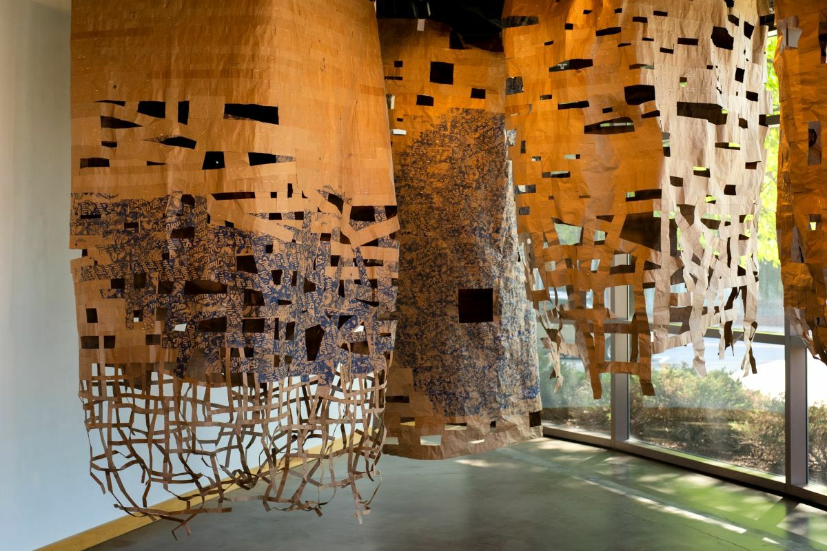 Large-scale suspended paper installation composed of layered brown and printed paper sheets, cut and woven into intricate, lattice-like forms. The hanging structures cascade from the ceiling in cylindrical shapes, with torn openings and dense collage areas. Natural light from floor-to-ceiling windows filters through the cutouts, casting soft shadows across the gallery floor.