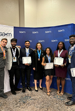 A group of six students and a faculty member posing for a photo, while holding awards, in front of a SAM banner.