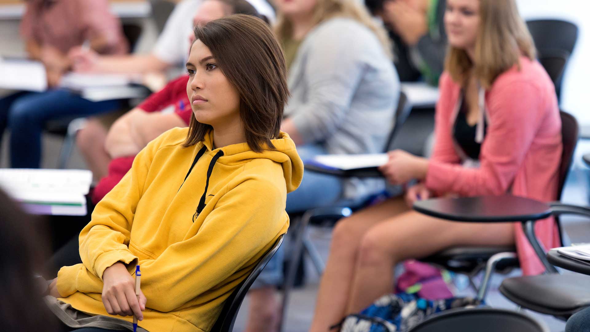 Students sitting in a classroom, one in a yellow hoodie, others blurred in the background.