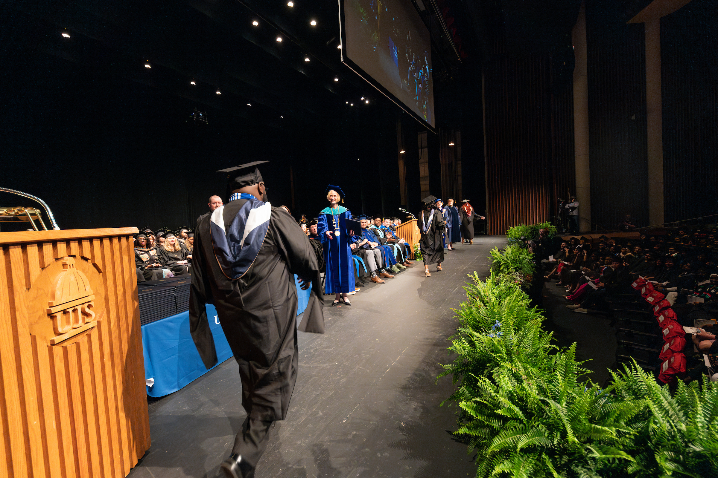 A student dressed in regalia walks across the Sangamon Auditorium stage to accept their diploma from the chancellor.