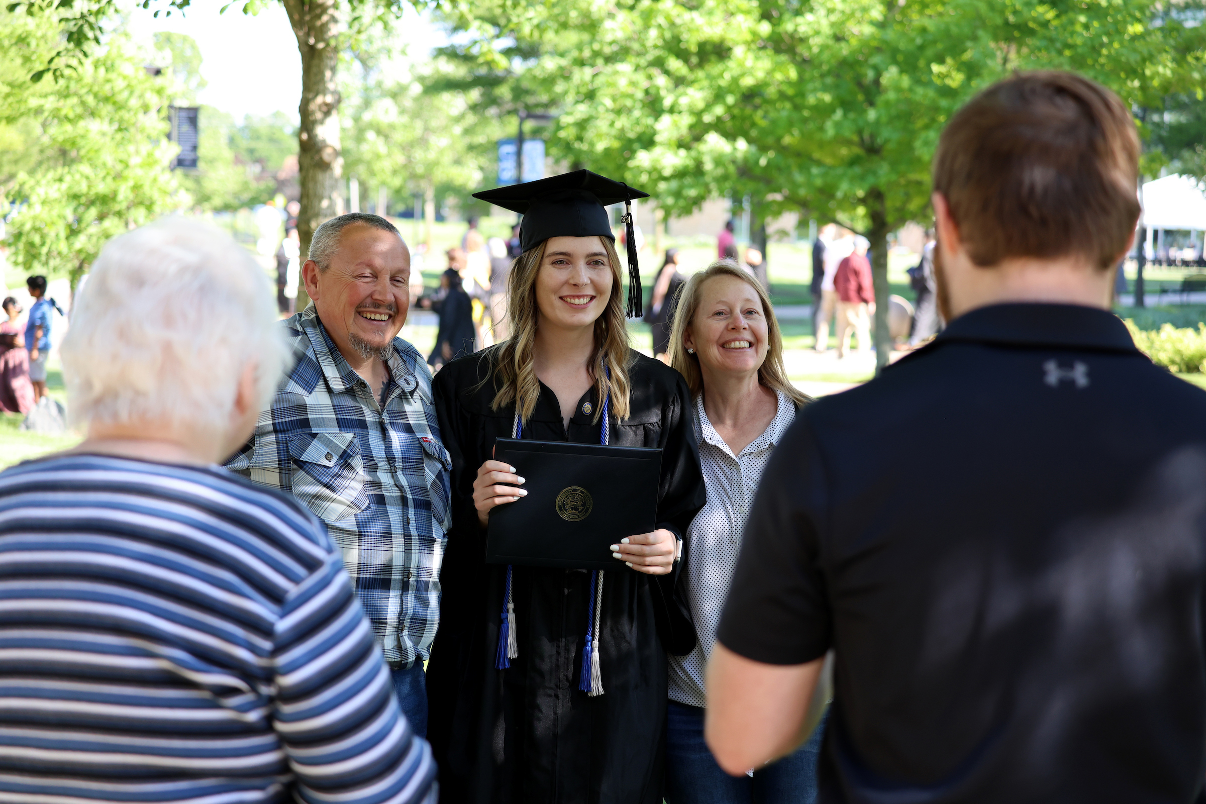 A woman in regalia stands flanked by a man and a woman, posing for a photo with her diploma cover.