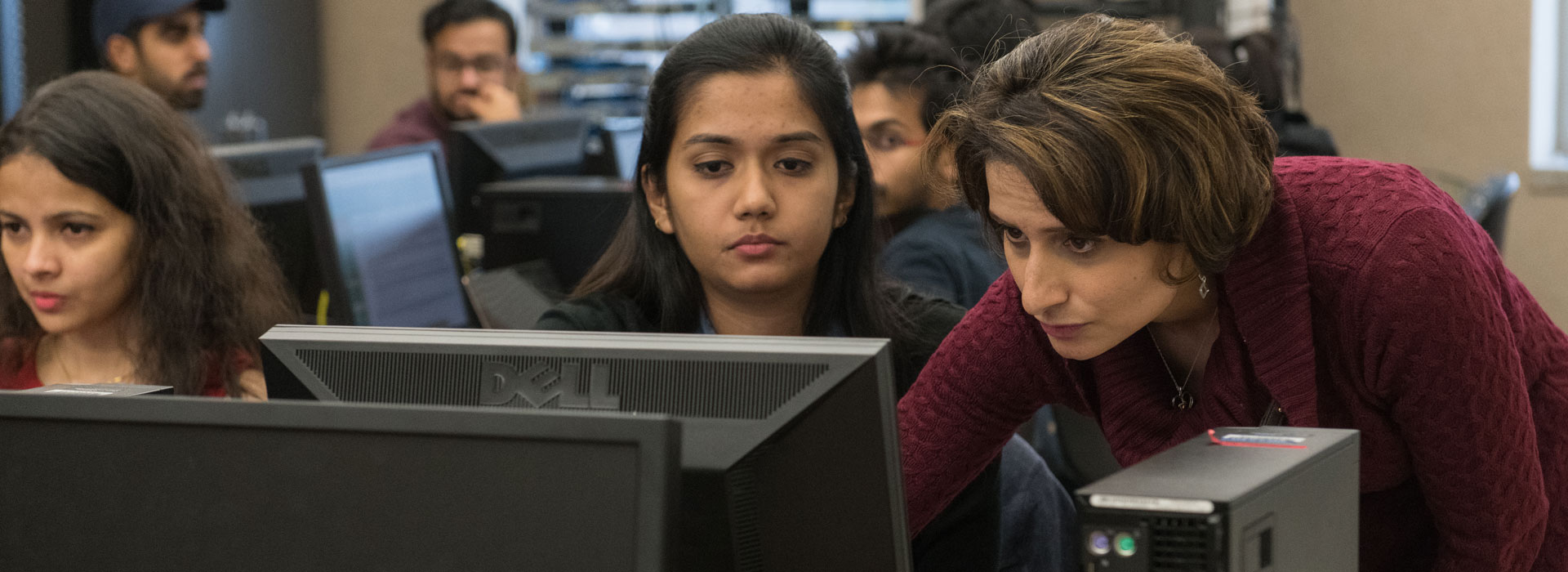 a professor standing next to a student at a computer desk showing the student something on a computer