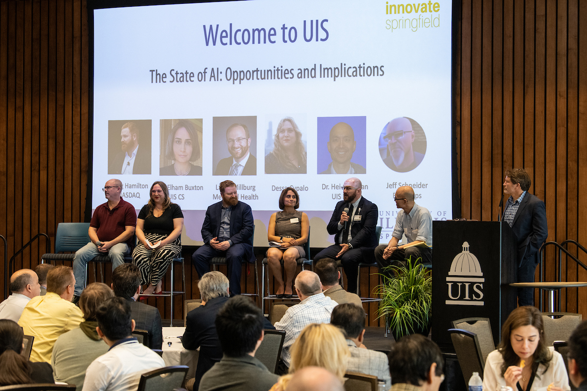 A panel of speakers sit in front of a screen projecting the name of their event: The State of AI: Opportunities and Implications.