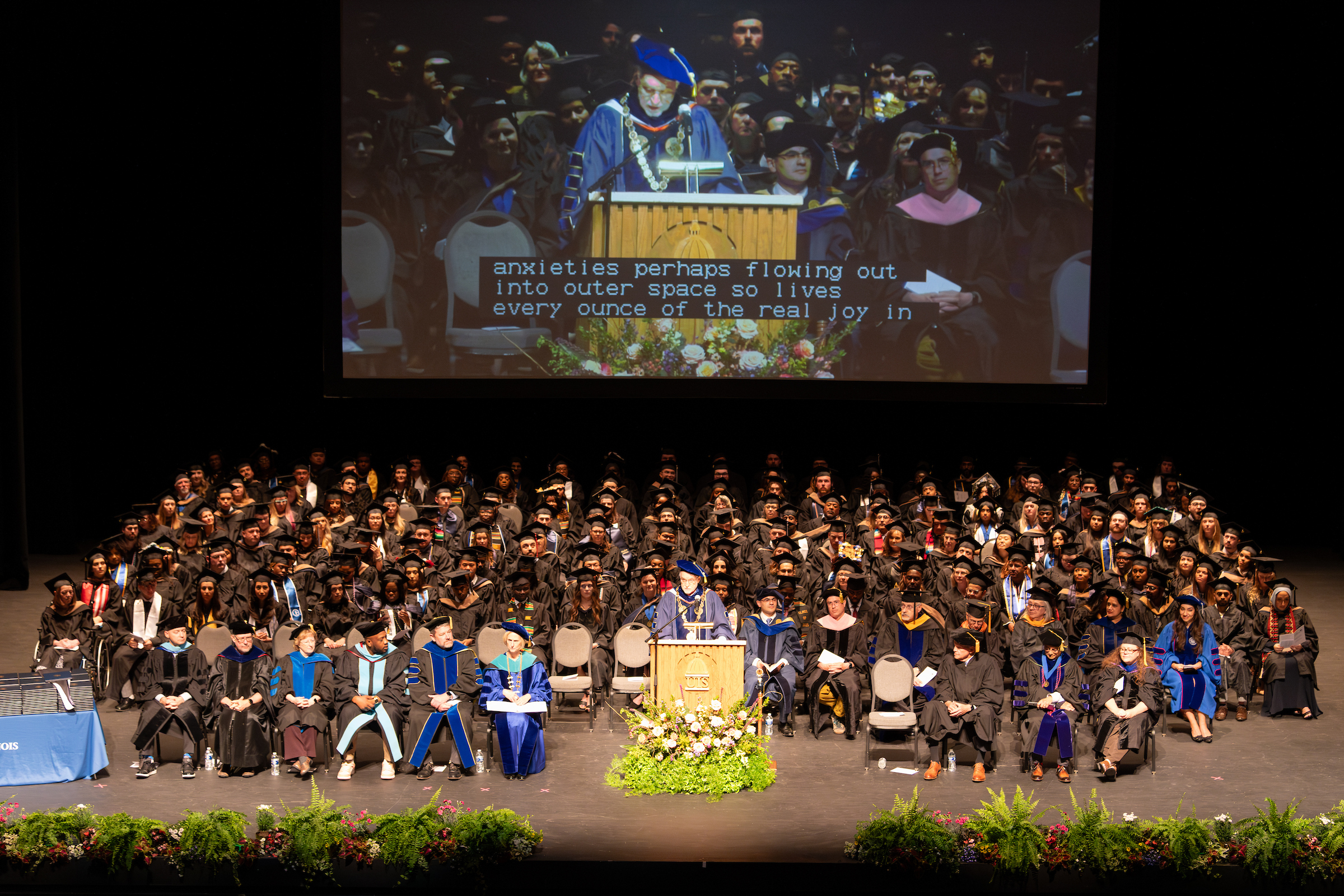A large group of CBM Students and faculty sit on stage facing the audience at a commencement ceremony.