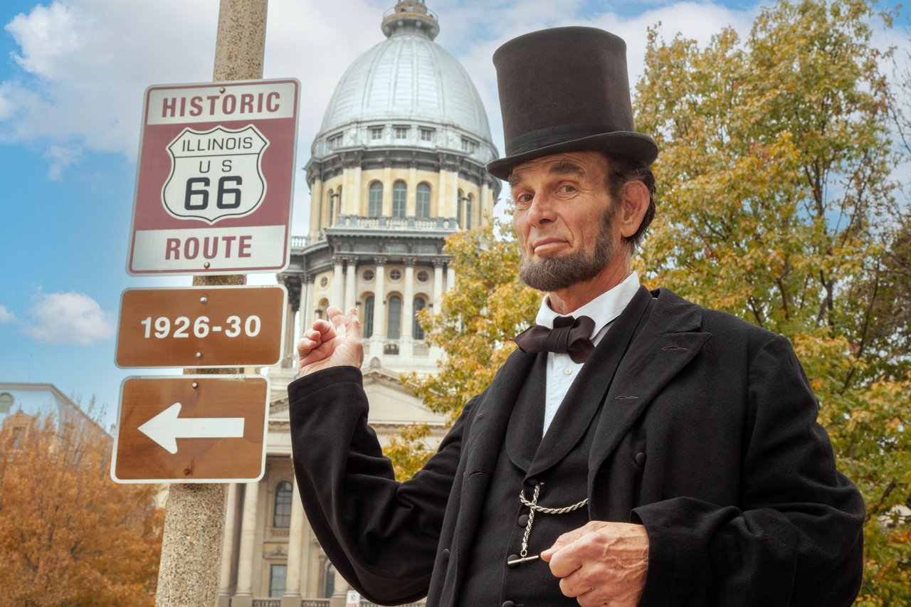 Abe Lincoln stands in front of the Illinois State Capitol and points at a historic Route 66 sign.