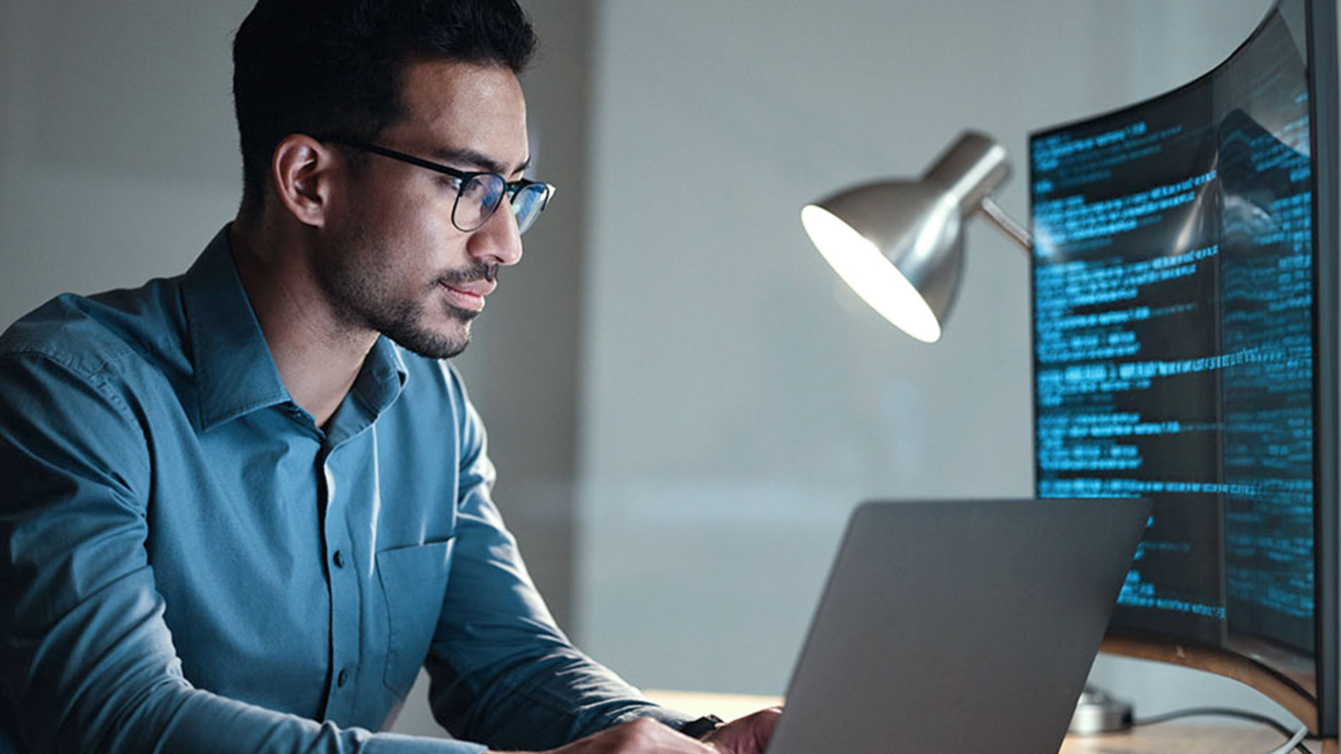 Man coding at night, illuminated by screen and lamp.