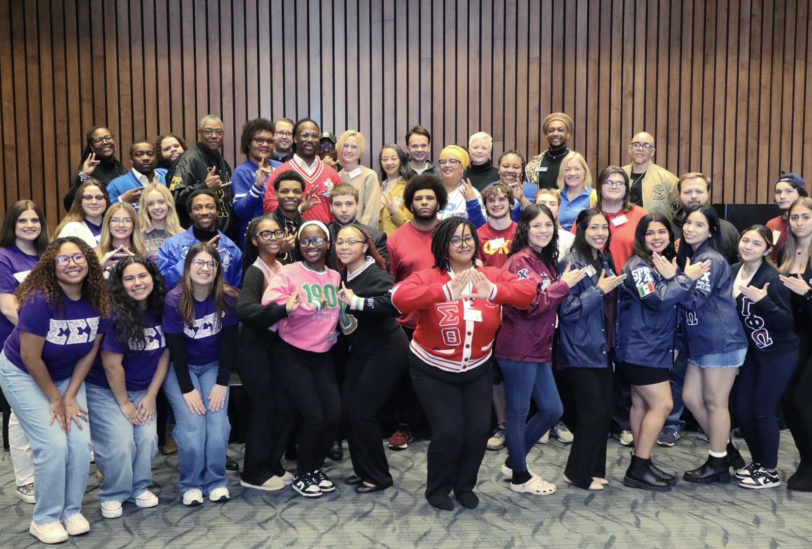 A large group of fraternity and sorority members posing together in front of a wood-paneled wall.