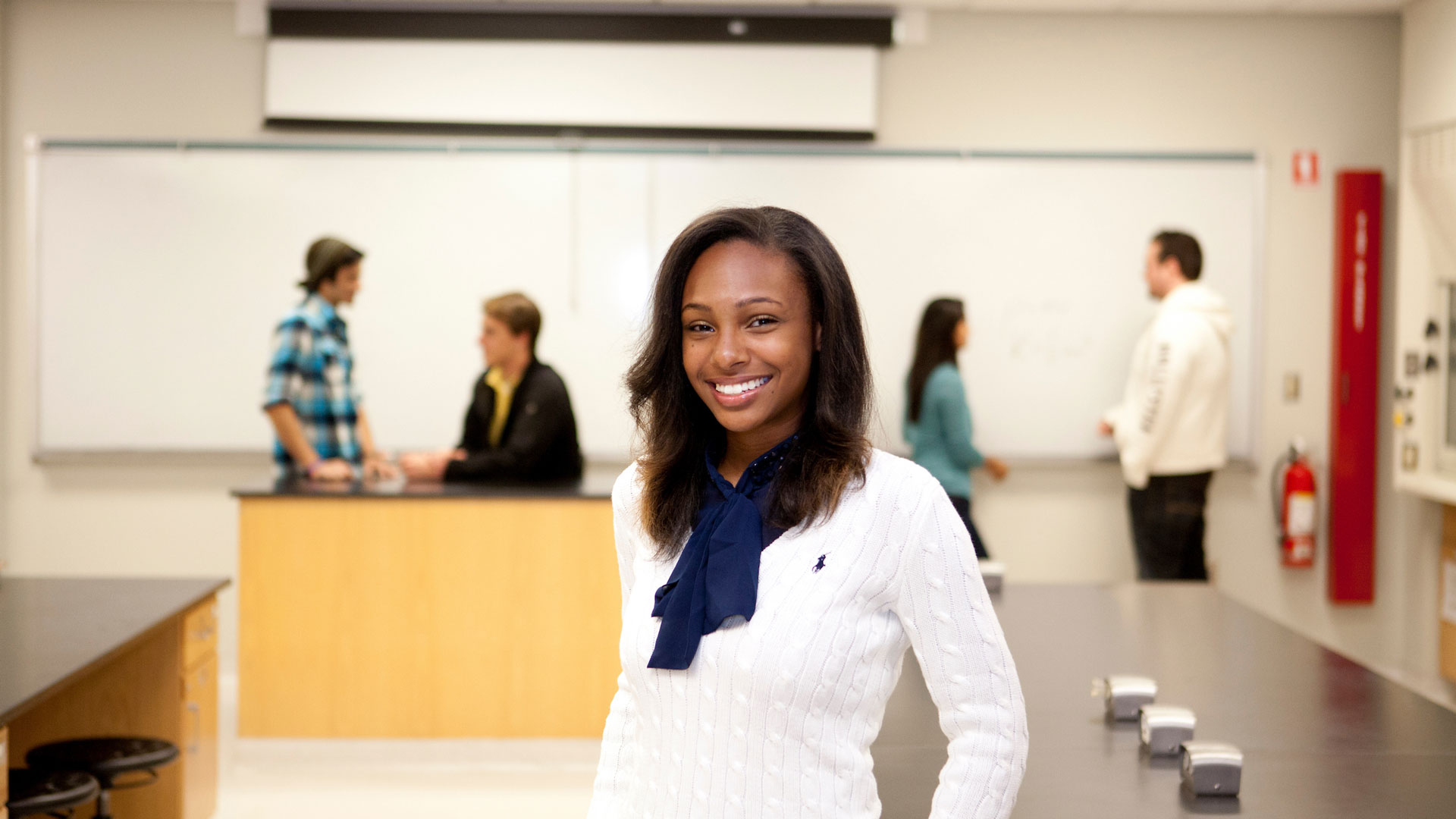 a female student smiling in a classroom setting with a professor and students in the background