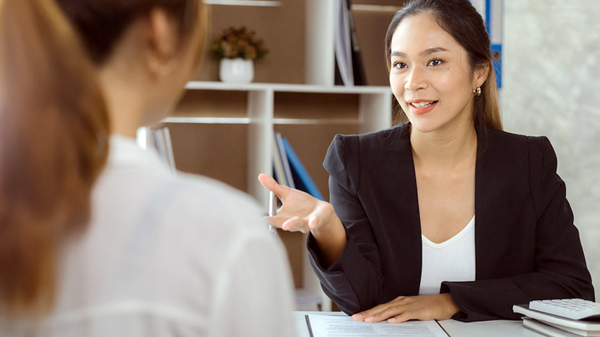Businesswoman in a black blazer talking to another person in an office setting.