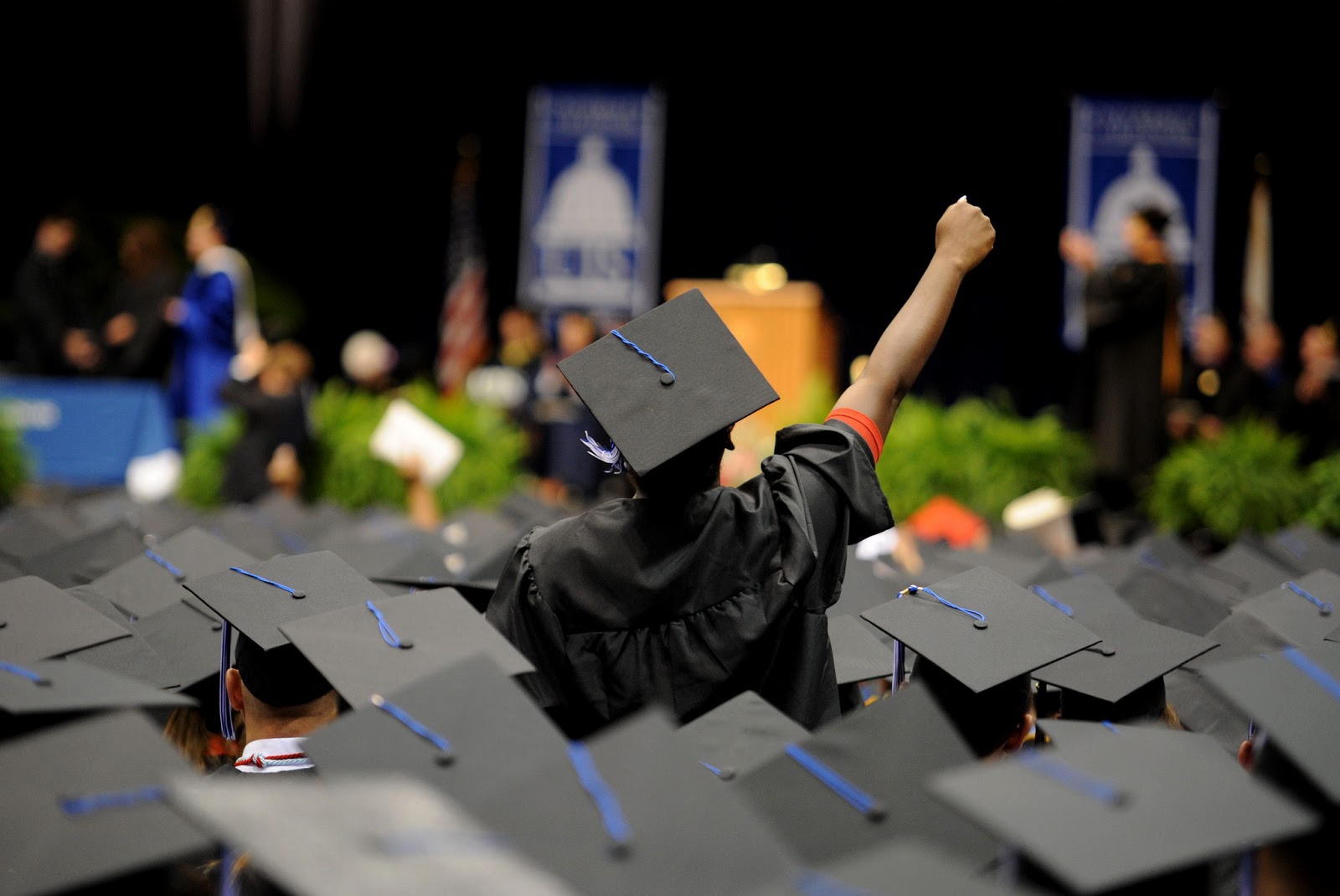 graduate standing up in the middle of a seated crowd of graduates pumping their fist in the air in triumph