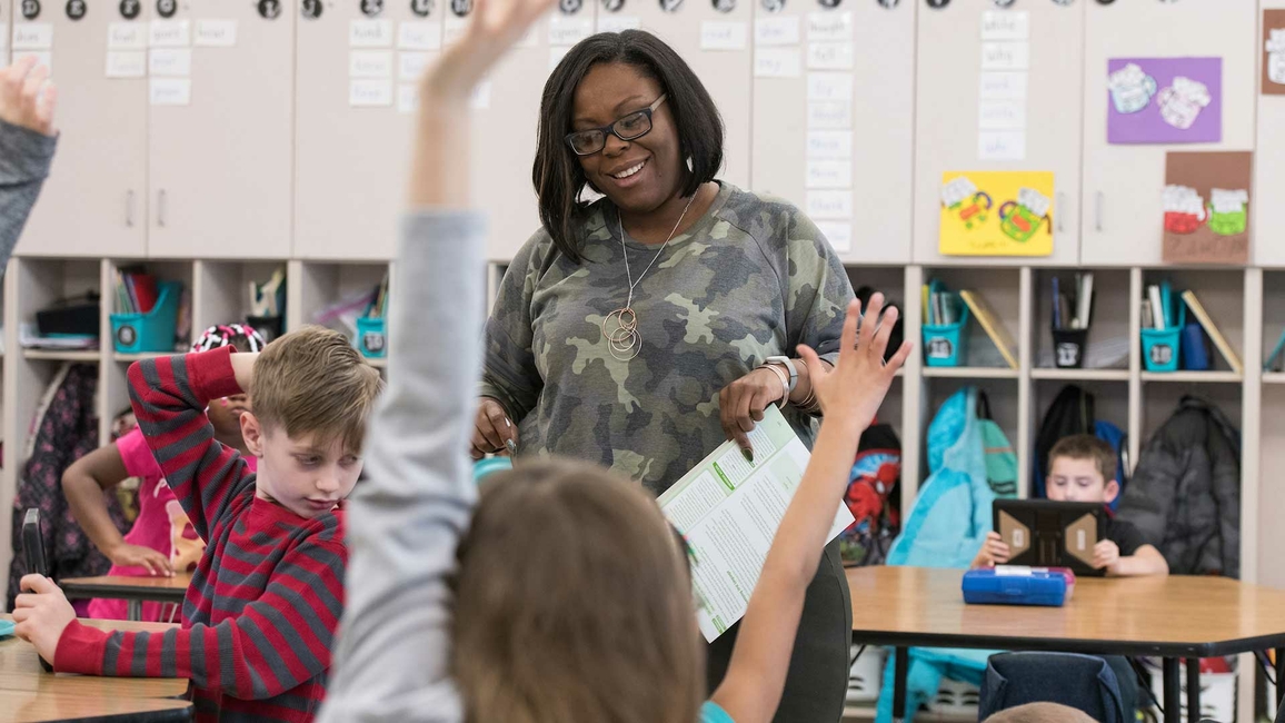 A teacher in a classroom smiling while students raise their hands.