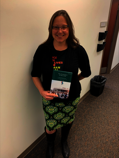 Dr. Kristi Barnwell, a white woman in a black sweater and green dress, smiling and holding a copy of her new book. 