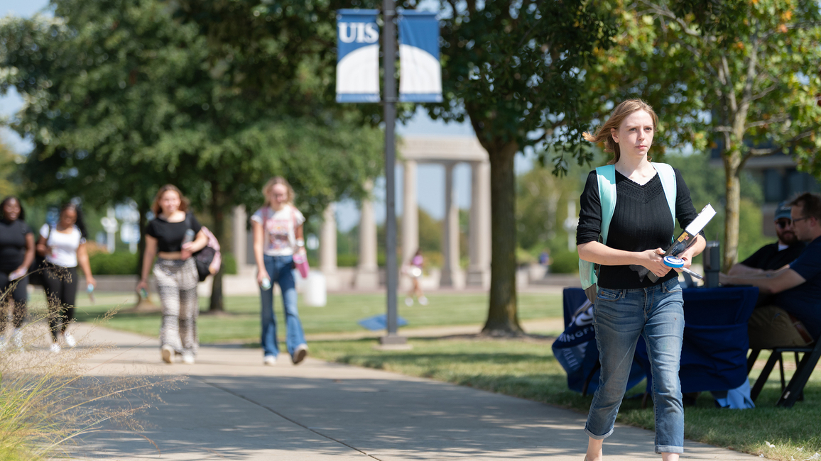 A student carrying books walking on a sidewalk with the colonnade in the background.