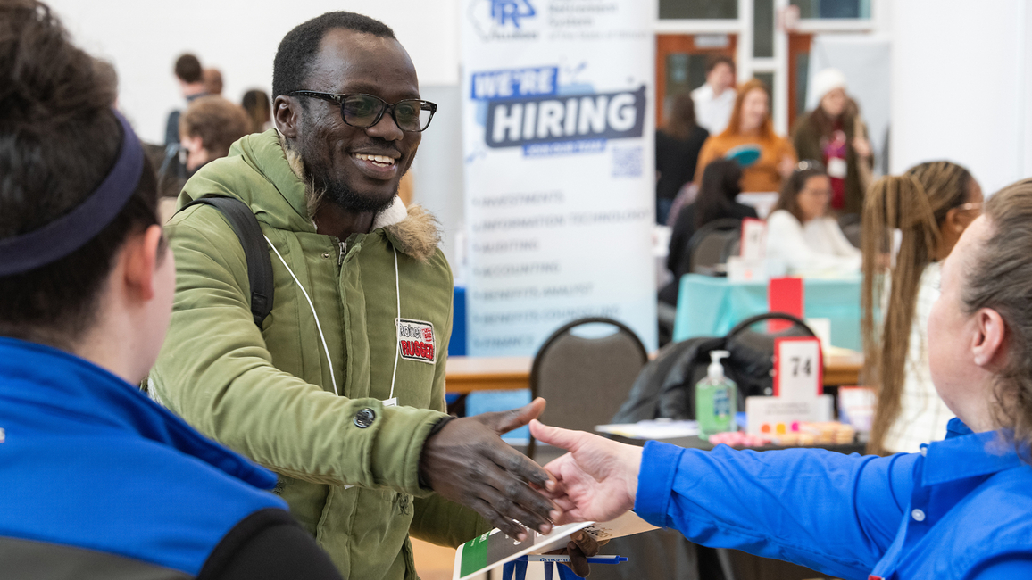 A smiling job fair attendee shakes hands with a recruiter at a table with a "We're Hiring" sign in the background.