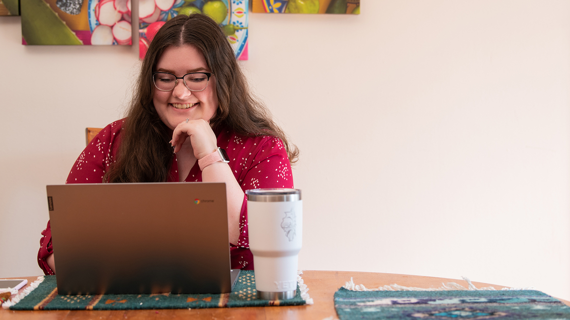 A person in glasses, wearing a red shirt, smiles while working on a laptop at a table with a white travel mug nearby.