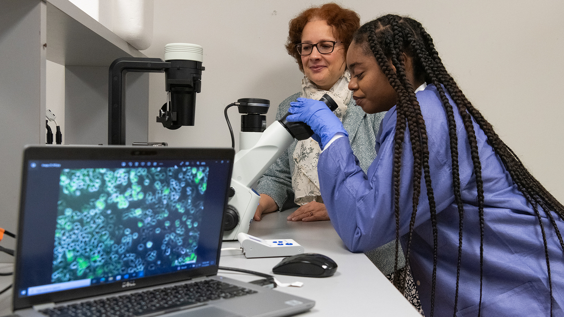 Student using a microscope in a lab while a professor observes, with cell images displayed on a nearby laptop screen.