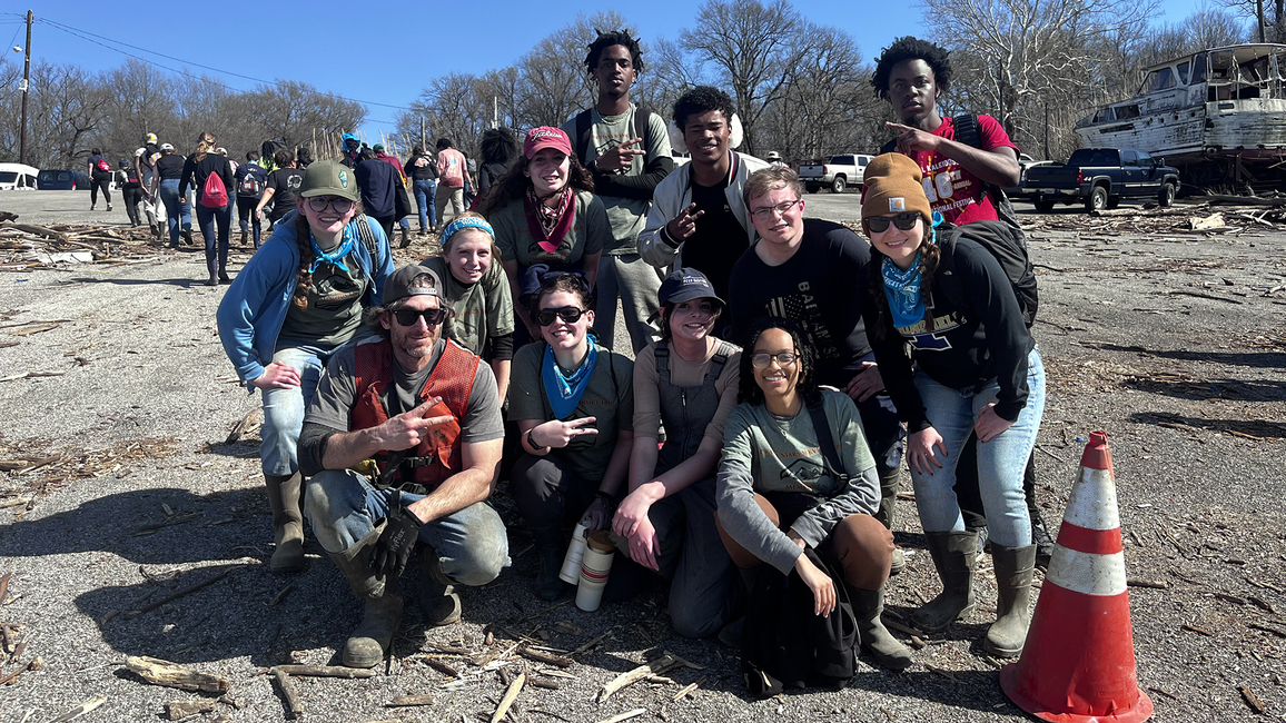 A group of volunteers poses for a photo during an outdoor cleanup event, wearing work gloves, boots, and bandanas. A damaged boat and debris are visible in the background.