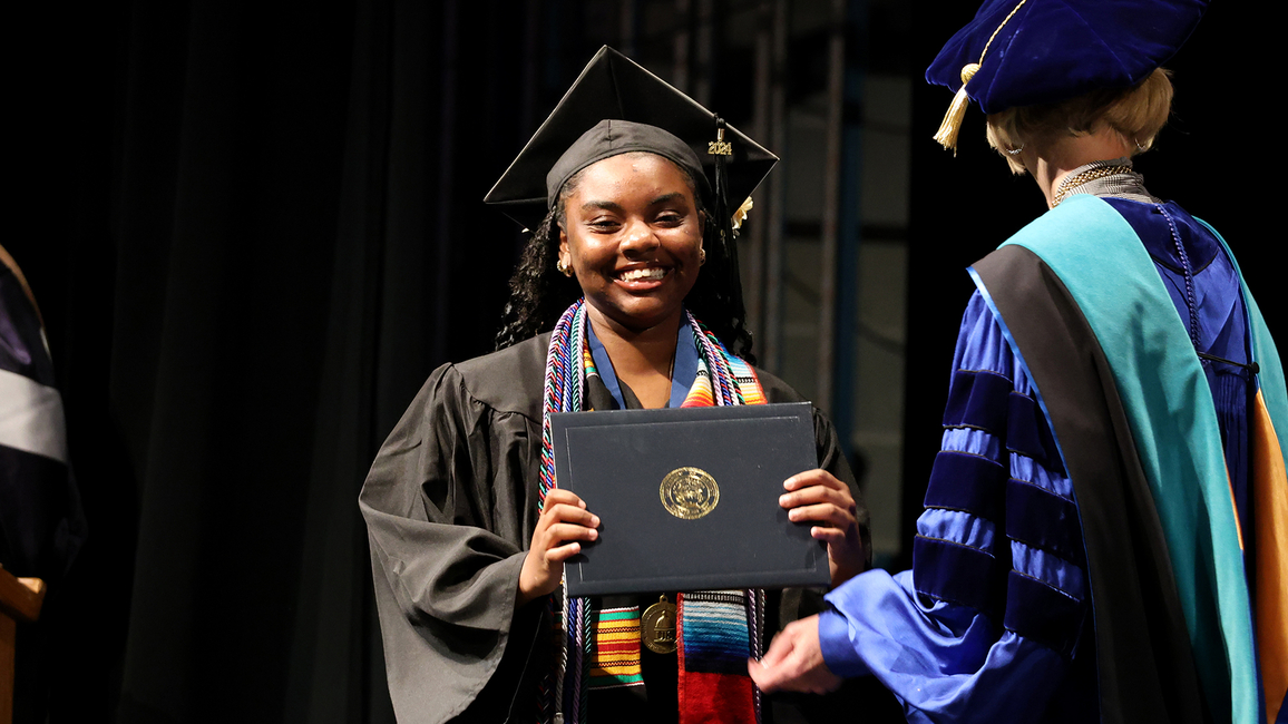 A UIS graduate smiles while holding her diploma on stage during the commencement ceremony, wearing a cap and gown.