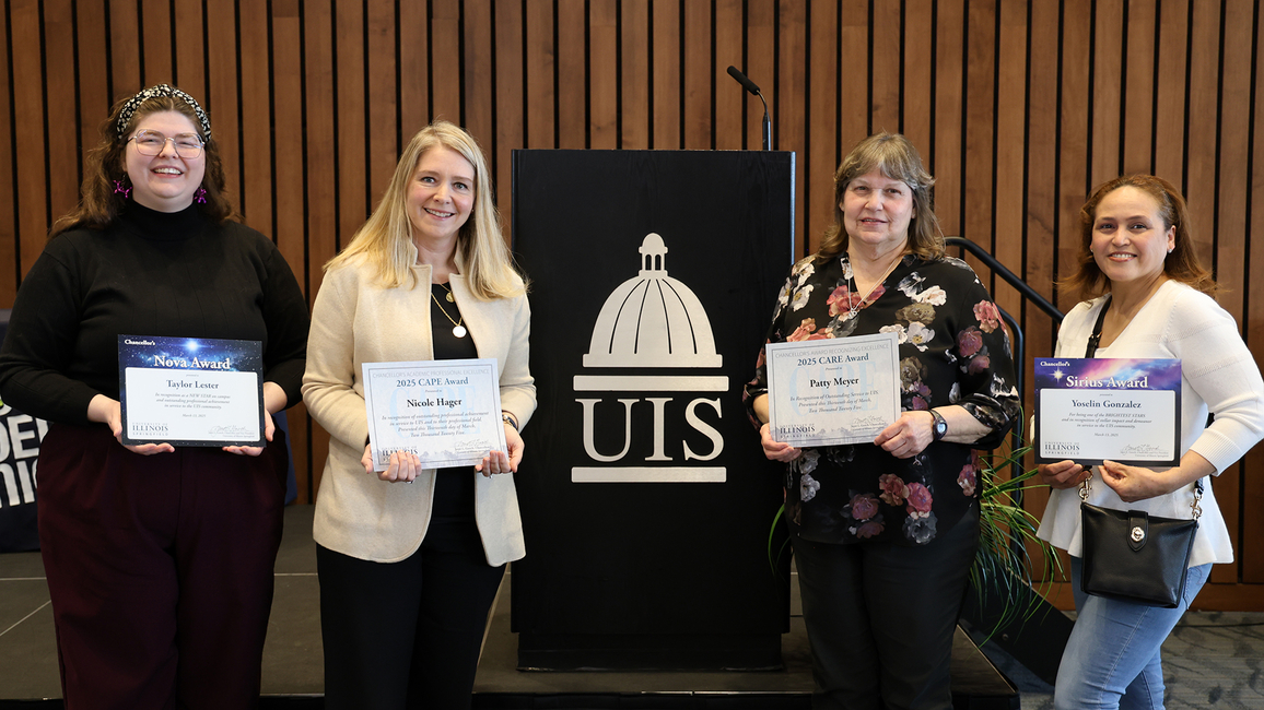 Four award recipients pose with their certificates in front of a UIS podium at a recognition event.