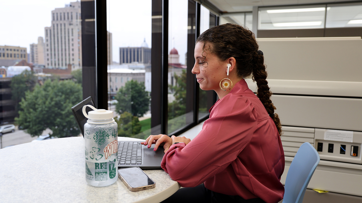 Young woman works on a laptop at a high-rise office window with earbuds, water bottle, and phone nearby.