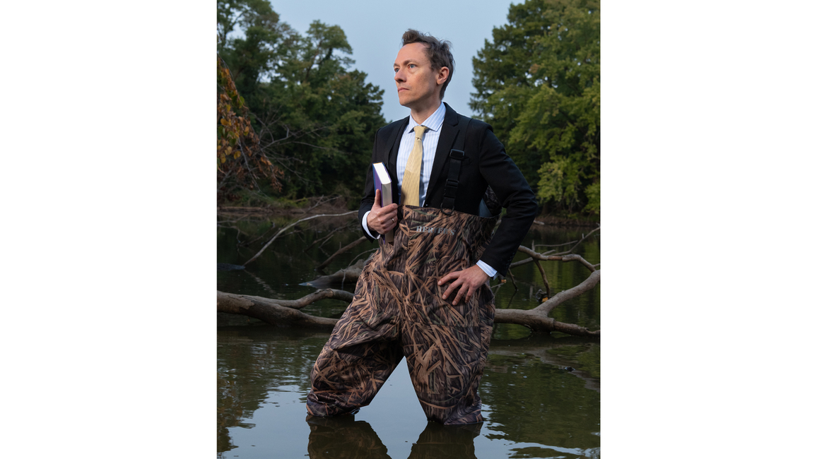 Brandon Derman stands in water wearing waders and a suit, holding a book, with trees in the background.