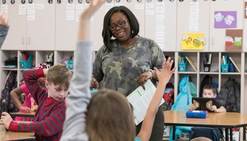 A teacher in a classroom smiling while students raise their hands.