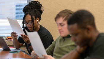 Three students sitting at a desk in a classroom reading pieces of paper. 