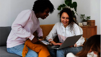 Two young women collaborating on a project while sitting on a couch in a cozy, well-decorated living room. One woman is holding a clipboard and leaning in, smiling as she talks with her friend, who is focused on her laptop. They both look engaged and happy, creating a warm and friendly atmosphere.