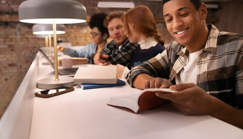 A group of students studying together at a communal table in a modern library. The student in the foreground is smiling and reading, while others are engaged in discussions and using laptops