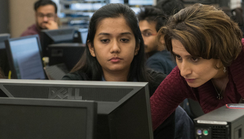 a professor standing next to a student at a computer desk showing the student something on a computer