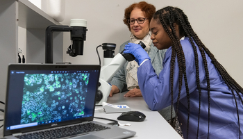 Student using a microscope in a lab while a professor observes, with cell images displayed on a nearby laptop screen.