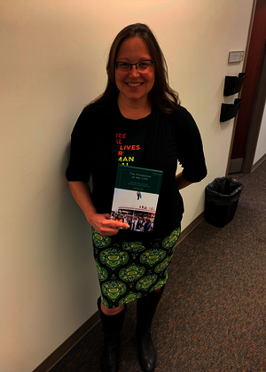 Photo of Dr. Kristi Barnwell, a white woman in a black sweater and green dress, smiling as she holds a copy of her new book.