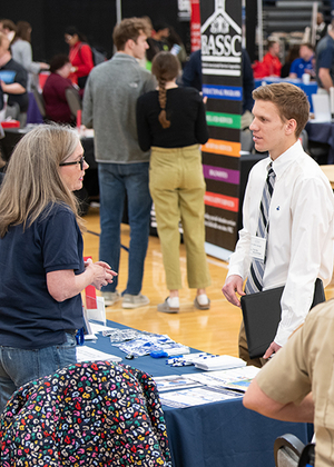 A job fair attendee wearing business attire speaks with a representative at an informational table.