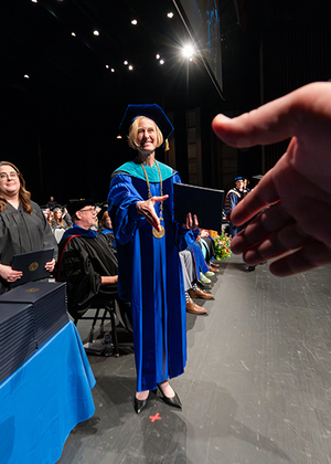 A university official in academic regalia extends a diploma and handshake to a graduate during a commencement ceremony.