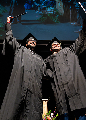 Two graduates in black caps and gowns celebrate on stage by raising their diplomas triumphantly, smiling as they stand side by side. Behind them, faculty members in academic regalia sit in rows and a large screen displays a projection of the moment above them.
