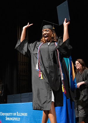 Graduating student celebrates on stage in cap and gown holding diploma at UIS Commencement as faculty applaud.