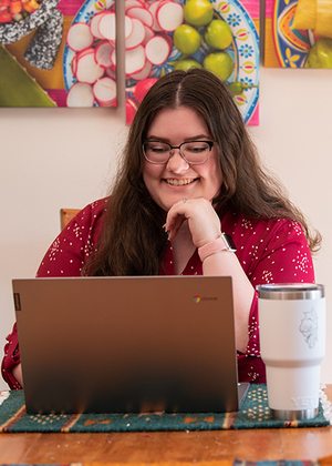 Student smiling while working on a laptop at a table with a tumbler, colorful art hanging on the wall behind her.
