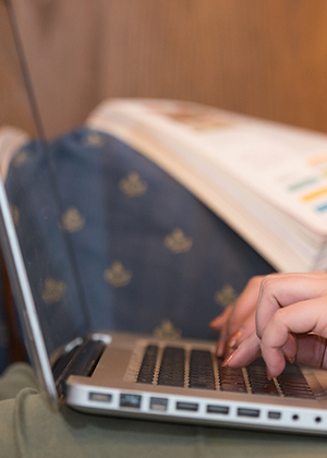 Close-up of a person typing on a laptop with an open textbook in the background.