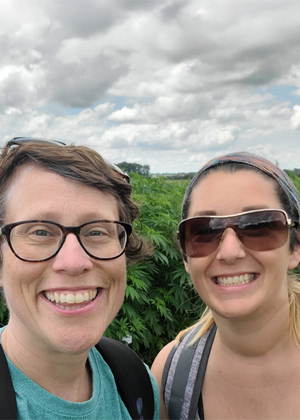 Two people smiling outdoors under a cloudy sky, surrounded by greenery.