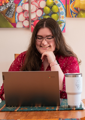 Woman smiling at a laptop with a large mug on the table; colorful art in the background.