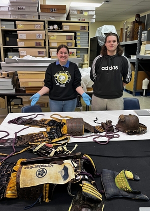Two people stand behind a table displaying historic Japanese armor. 