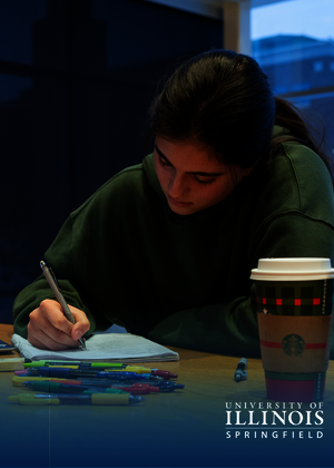 Student studying at a desk with a laptop and coffee cup, in dim lighting.
