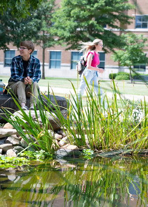 People sitting by a pond with greenery and a building in the background.