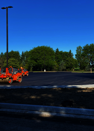 Freshly paved parking lot with orange traffic barrels and trees in the background.