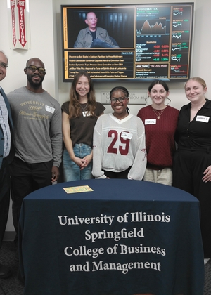 Group photo of MBA students and faculty at the Scoop & Socialize event, standing behind a University of Illinois Springfield College of Business and Management table.