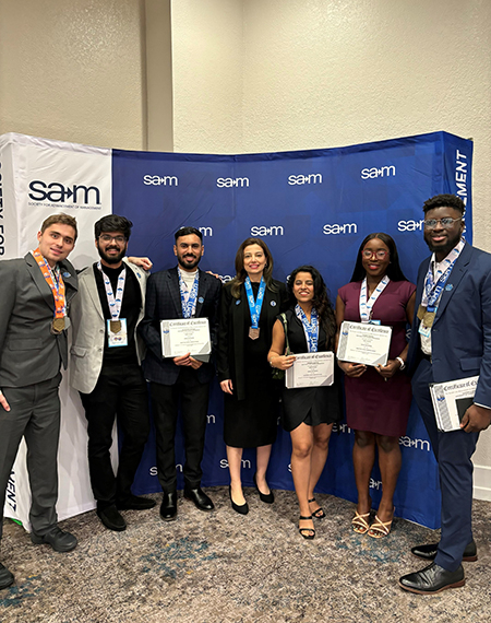 A group of six students and a faculty member posing for a photo, while holding awards, in front of a SAM banner.