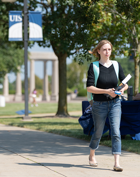 A student carrying books walking on a sidewalk with the colonnade in the background.