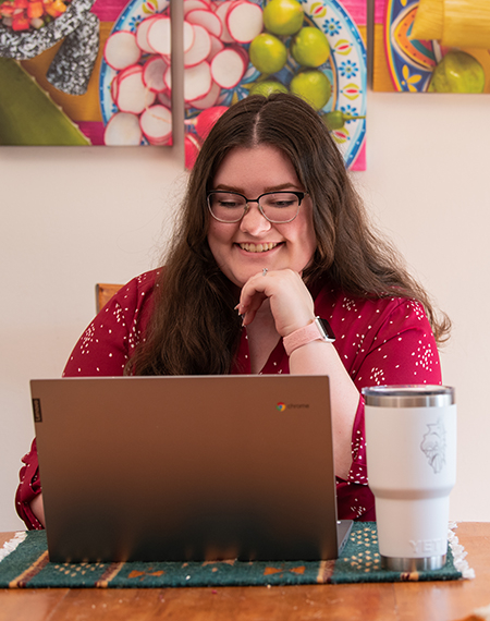 A person in glasses, wearing a red shirt, smiles while working on a laptop at a table with a white travel mug nearby.