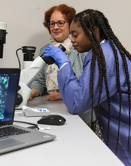 Student using a microscope in a lab while a professor observes, with cell images displayed on a nearby laptop screen.